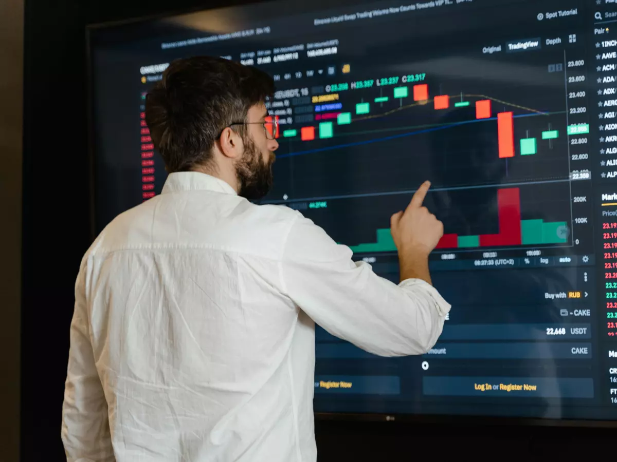 A man in a white shirt and glasses is looking at a large screen with a chart of stock prices. He is pointing with his right hand at the screen.