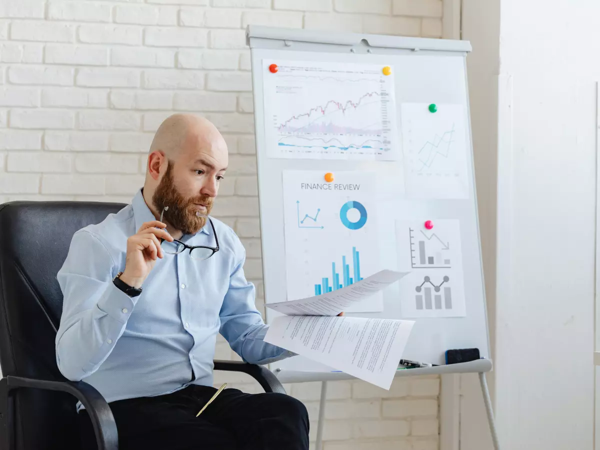 A man sits at a desk in an office, examining a document. He has a beard and is wearing a blue shirt, and the room is decorated with charts on a whiteboard in the background.