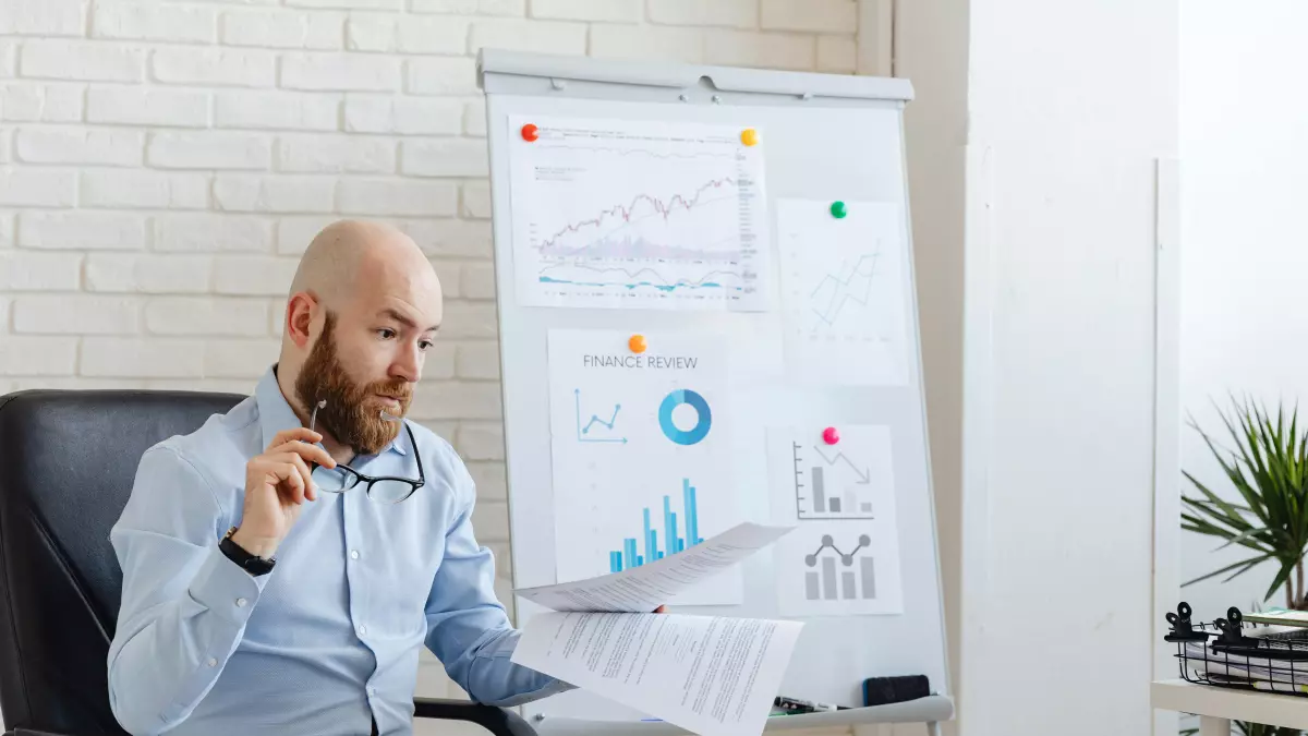 A man sits at a desk in an office, examining a document. He has a beard and is wearing a blue shirt, and the room is decorated with charts on a whiteboard in the background.