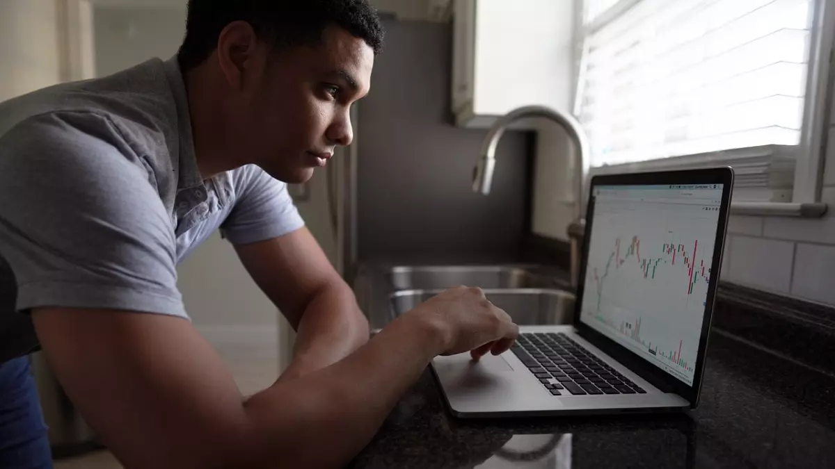 A young man is looking at a laptop screen. The laptop is on a counter. The man is wearing a gray shirt and jeans. The screen of the laptop shows a chart.