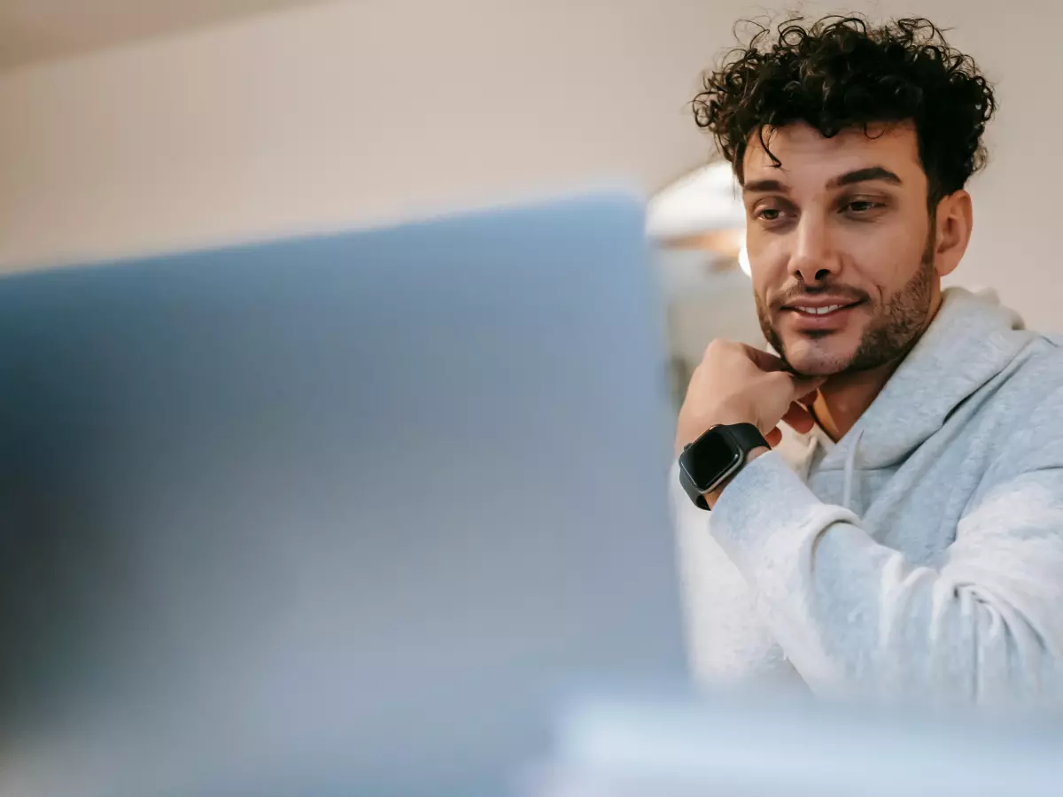 A man in a hoodie sits at a desk with a laptop and appears to be looking at a smartwatch on his left wrist.