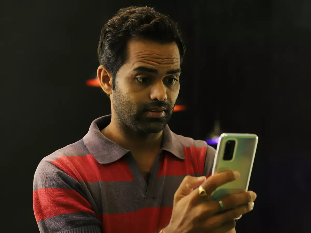 A man is holding a silver Samsung phone and looking at it. He is wearing a red and grey striped polo shirt. The man is standing in front of a dark background with blurred lights. 