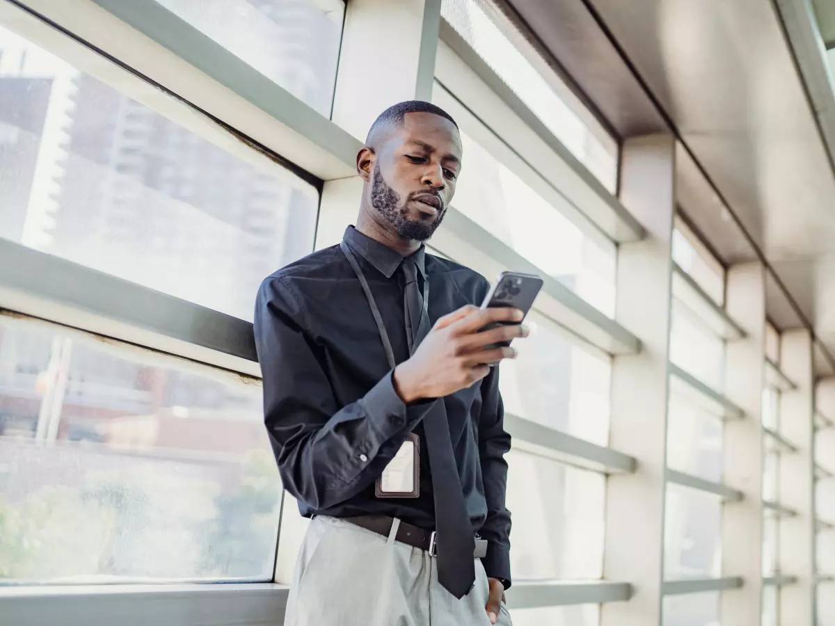 A man in a black shirt and gray pants stands in an office hallway and looks at his phone.