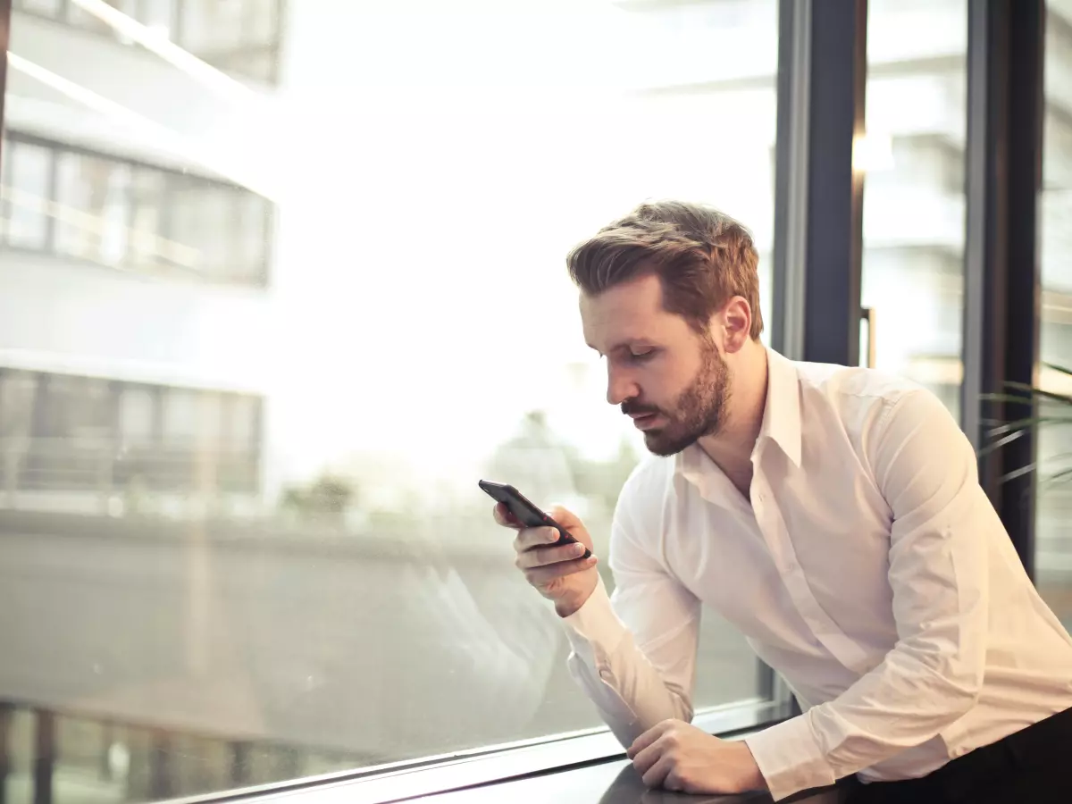 A man in a white shirt is looking at his phone while sitting by a window. He's in a modern setting with a view of the city.