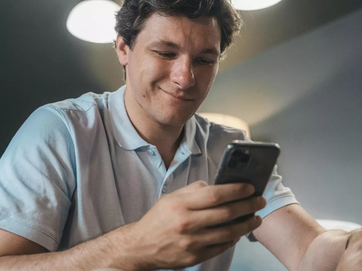 A man is smiling and looking at his phone, he is wearing a white shirt and sitting in a room with a bright light source above him.