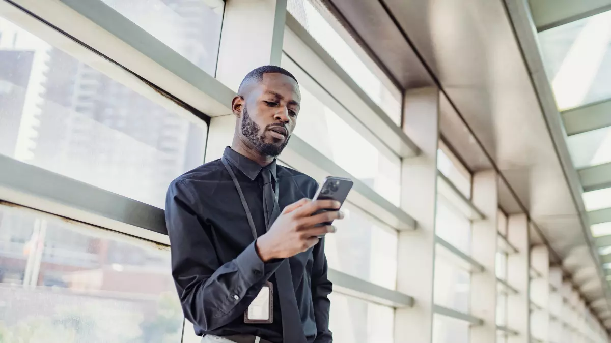 A man in a black shirt and gray pants stands in an office hallway and looks at his phone.