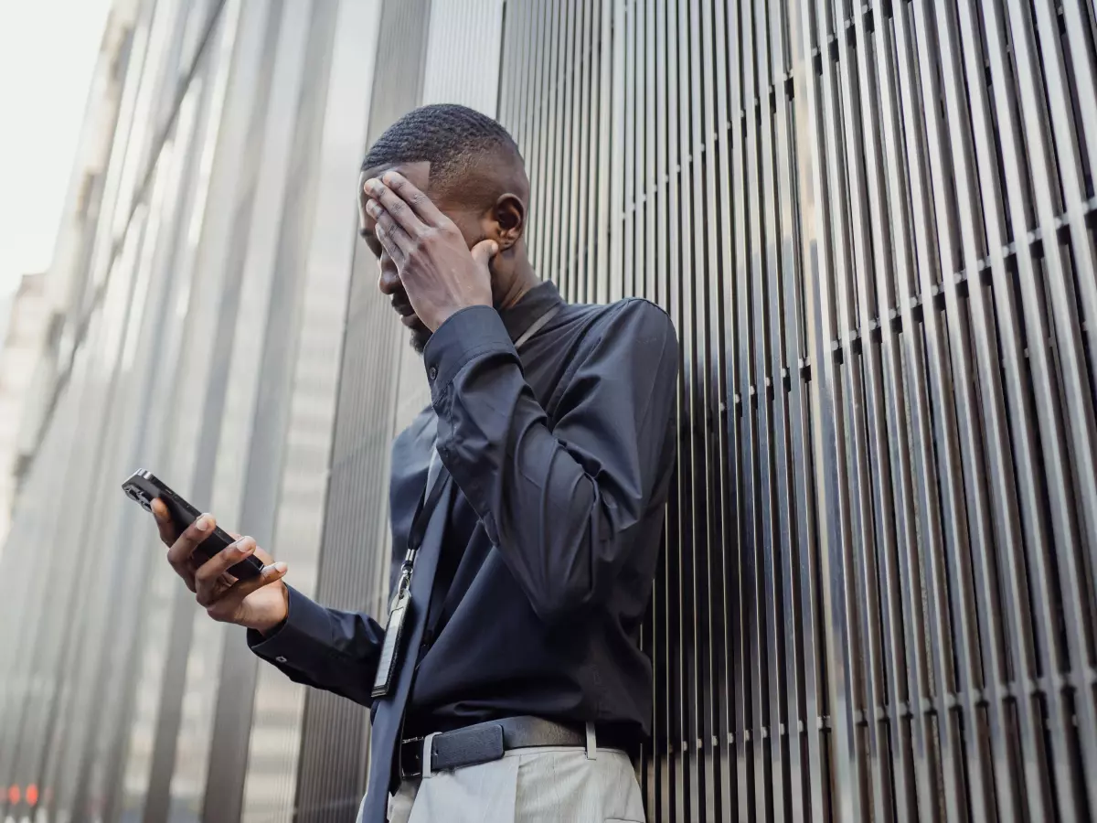 A young black man is standing in front of a building and holding his phone in his hand. His hand is over his face in surprise or shock.