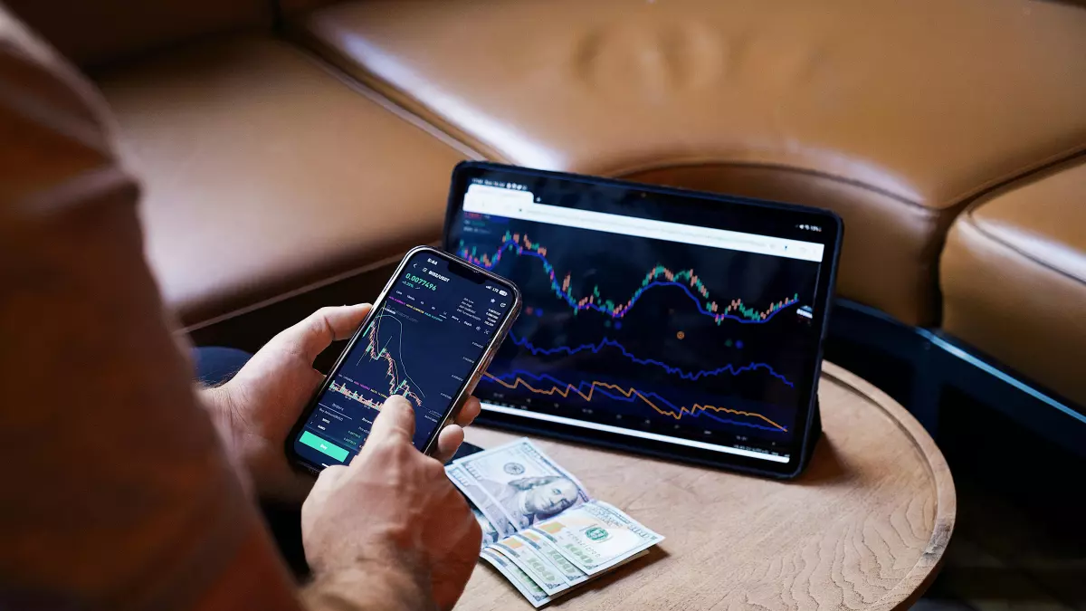 A person is sitting on a sofa and analyzing stock charts on their phone and laptop.