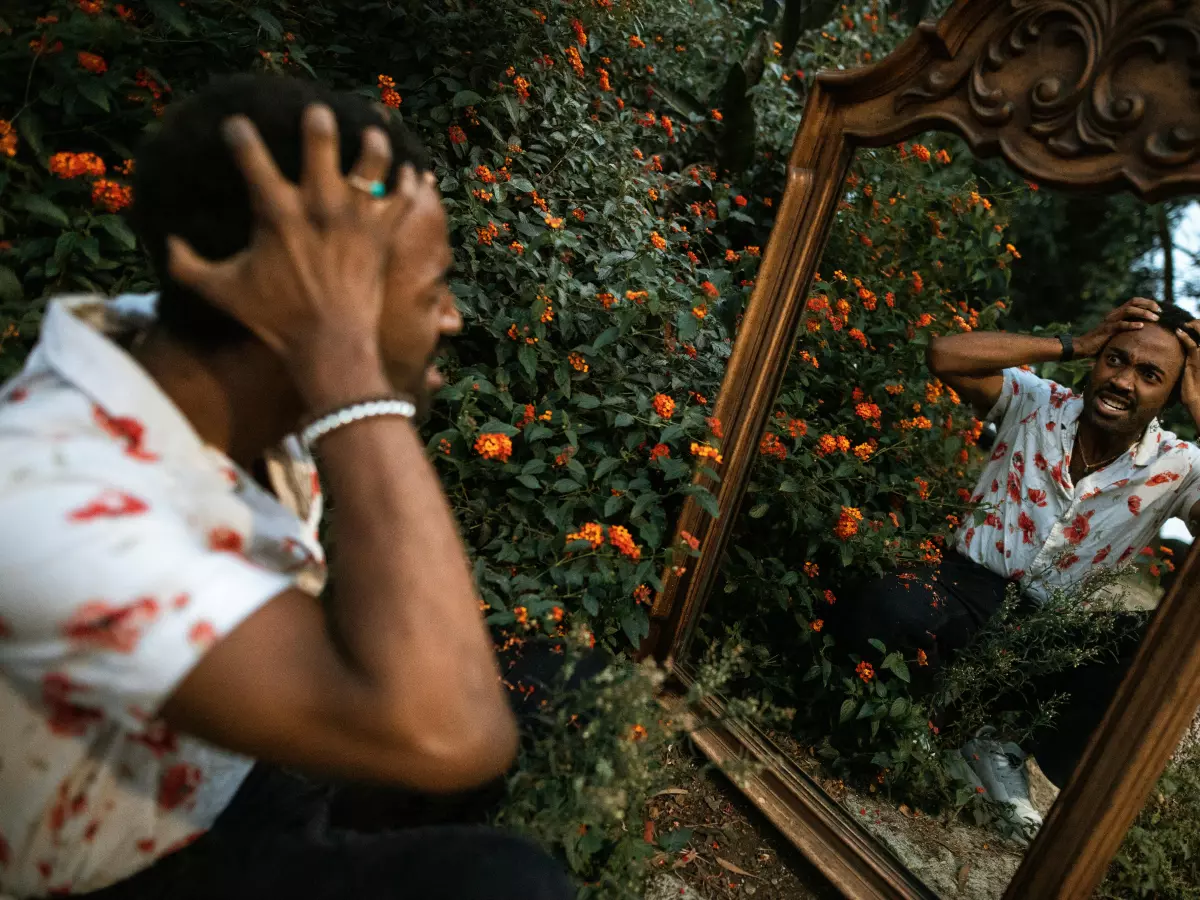 A man in a white shirt with red patterns is looking into a mirror and pulling his hair out in frustration.  The mirror is surrounded by green plants, with orange fruits in the background.