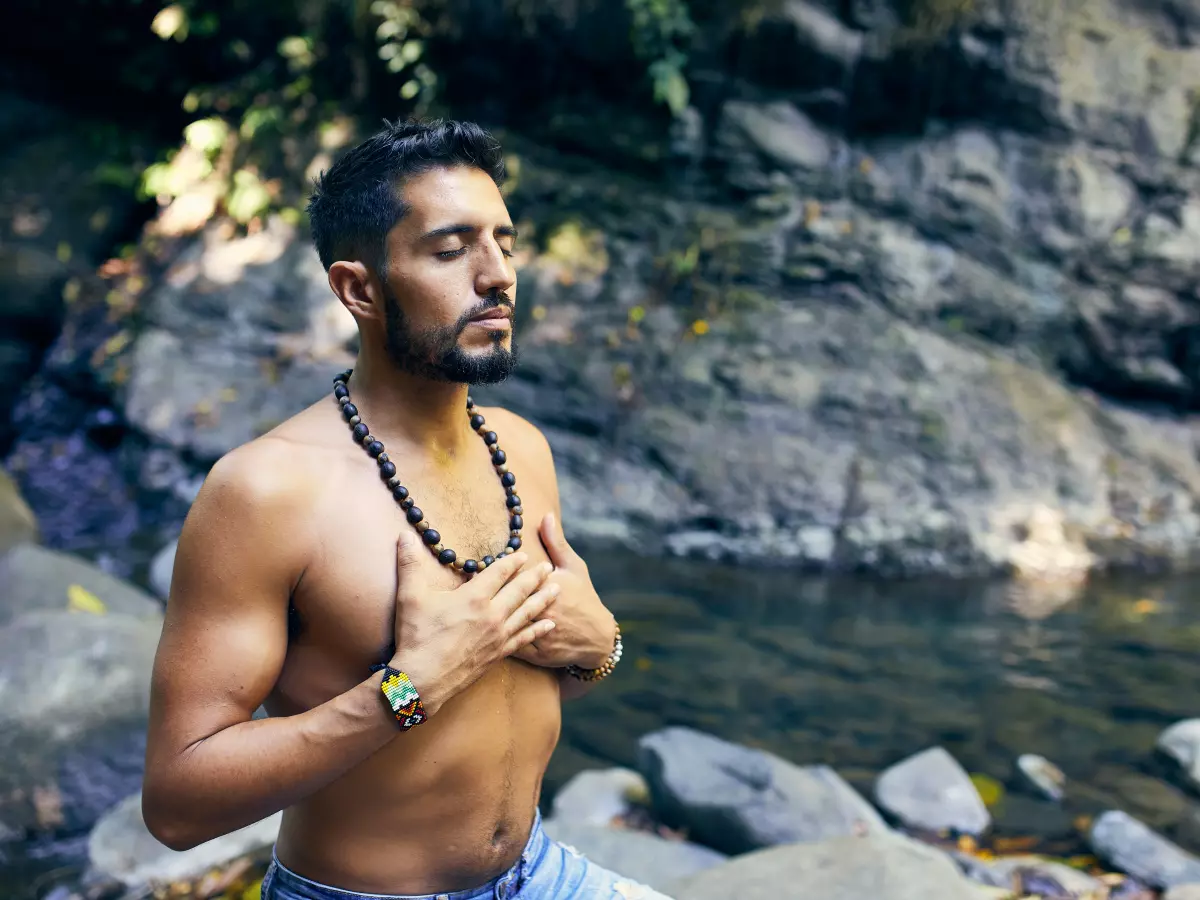 A man with a beard, shirtless, sits on rocks by a river, looking up. His hands are on his chest.