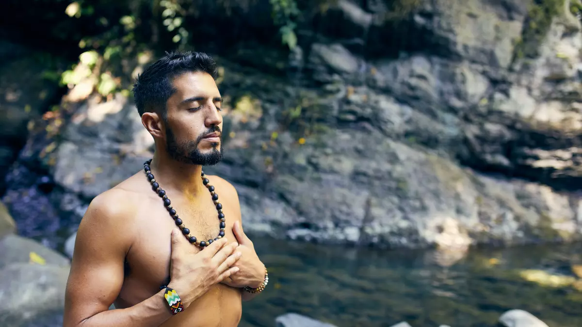 A man with a beard, shirtless, sits on rocks by a river, looking up. His hands are on his chest.