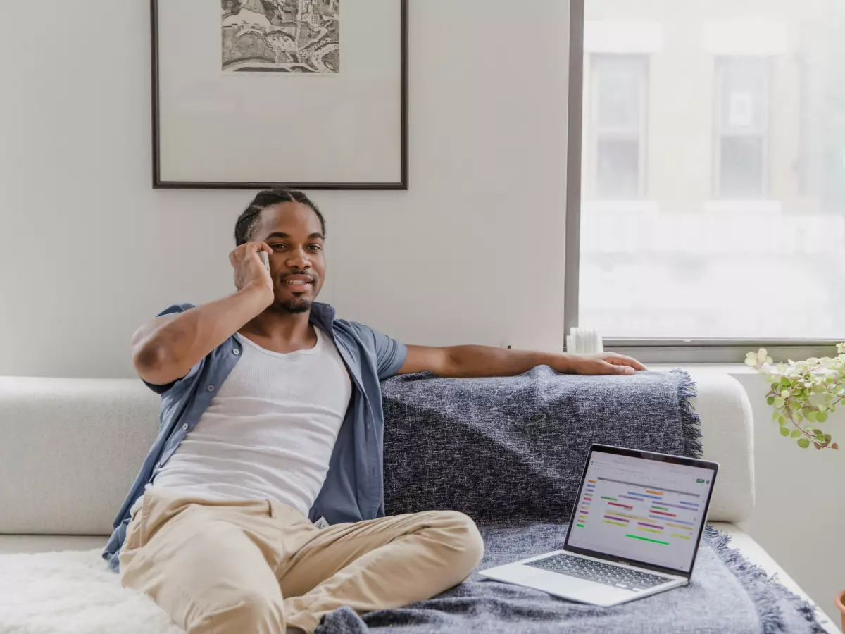 A man on the phone, sitting on a couch, looking at a laptop