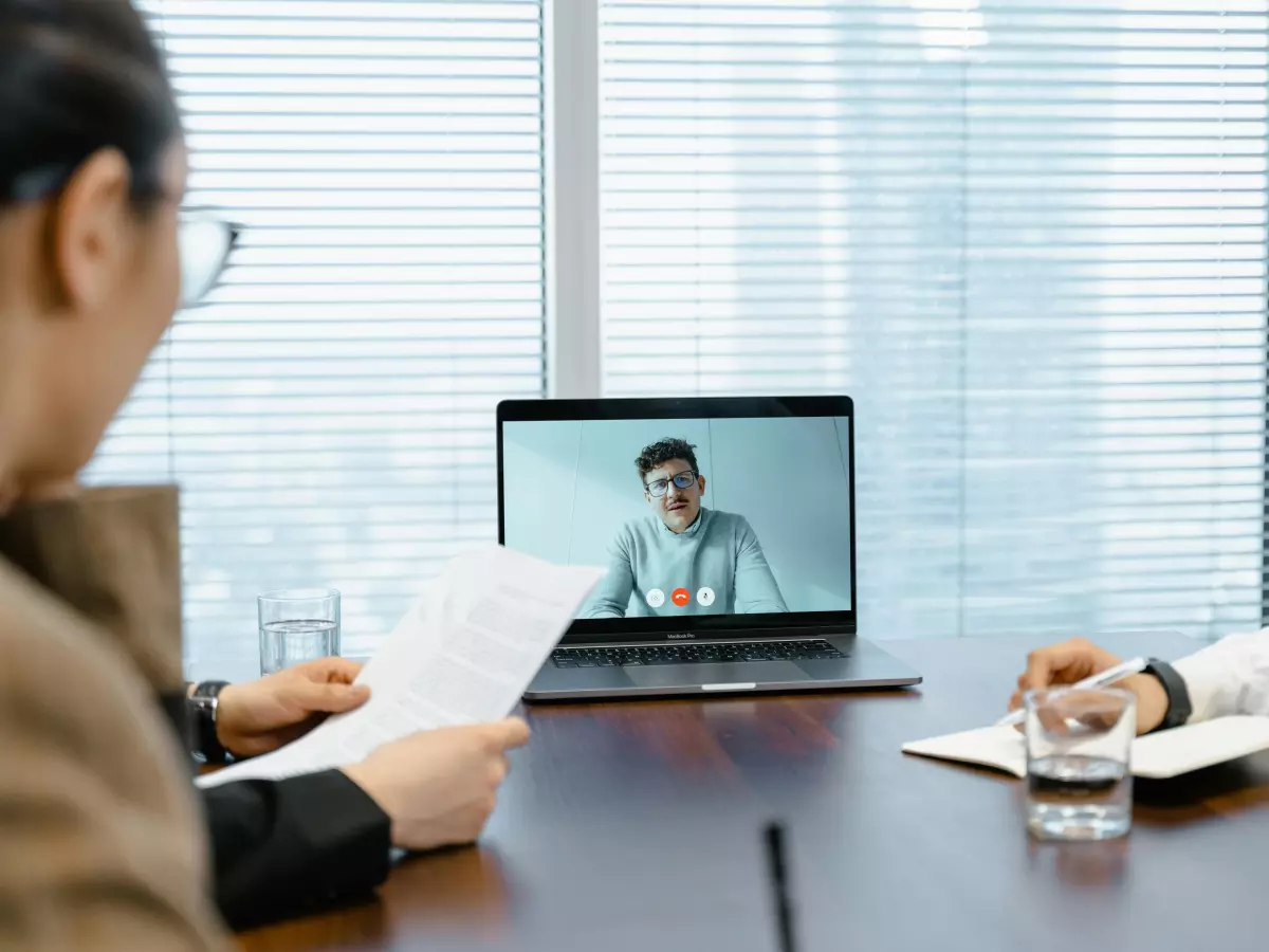 A professional interview scene. Two people are sitting at a desk, one with a paper and the other with a pen. The person on the laptop screen is smiling and speaking.