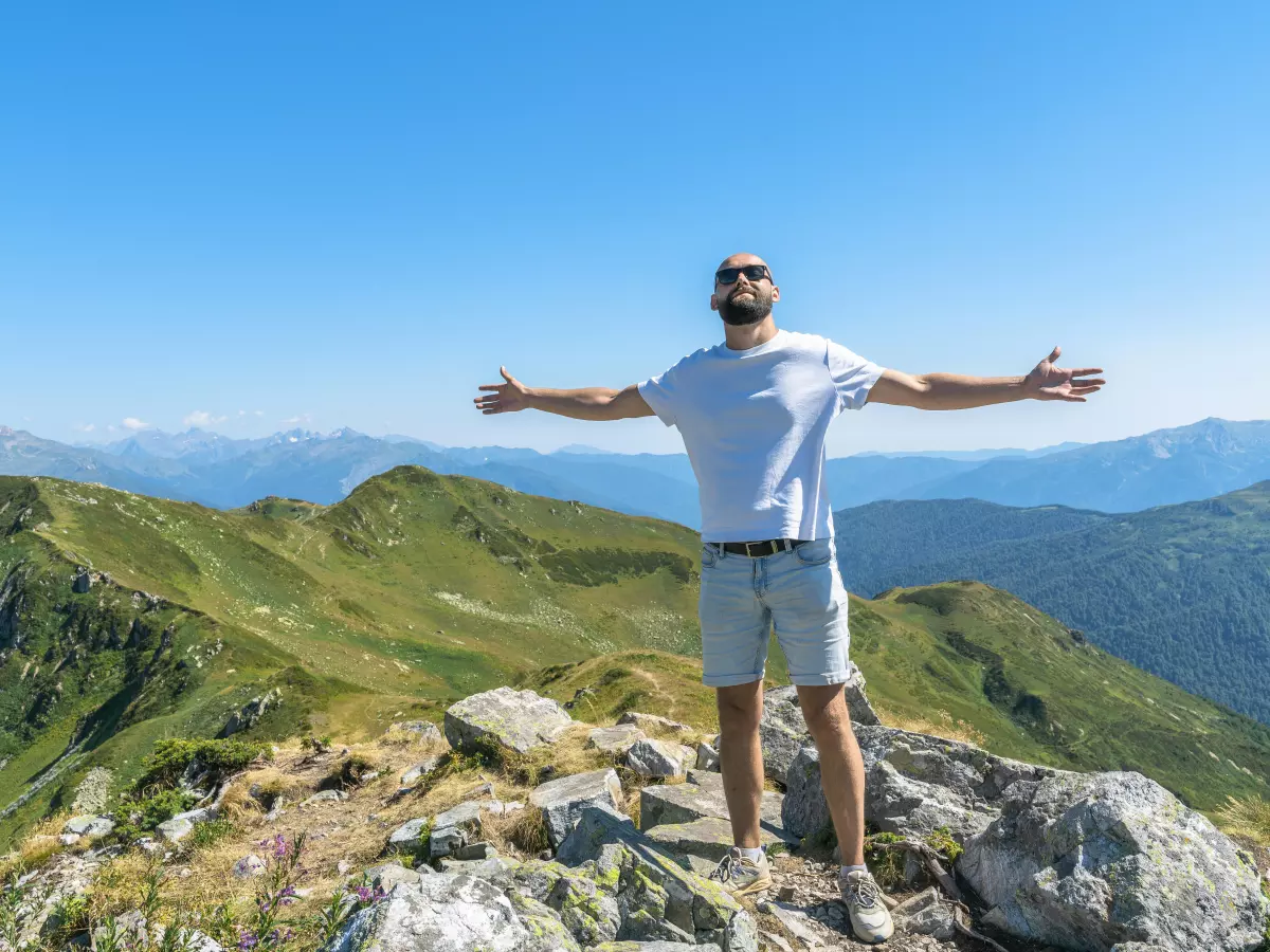 A man in a white t-shirt and shorts is standing on a mountaintop, looking out at the view with his arms outstretched. The mountains are covered in green grass and the sky is blue.