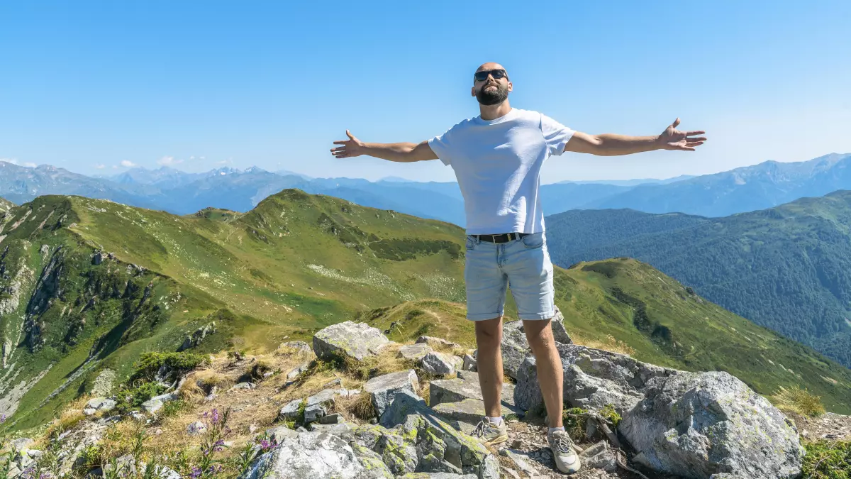 A man in a white t-shirt and shorts is standing on a mountaintop, looking out at the view with his arms outstretched. The mountains are covered in green grass and the sky is blue.