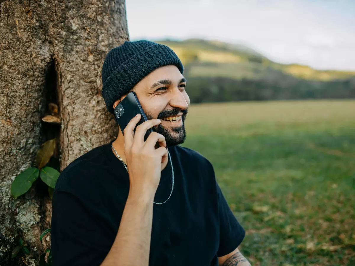 A young man with a beard is sitting on a grassy field with a tree behind him, he is smiling while talking on his phone.