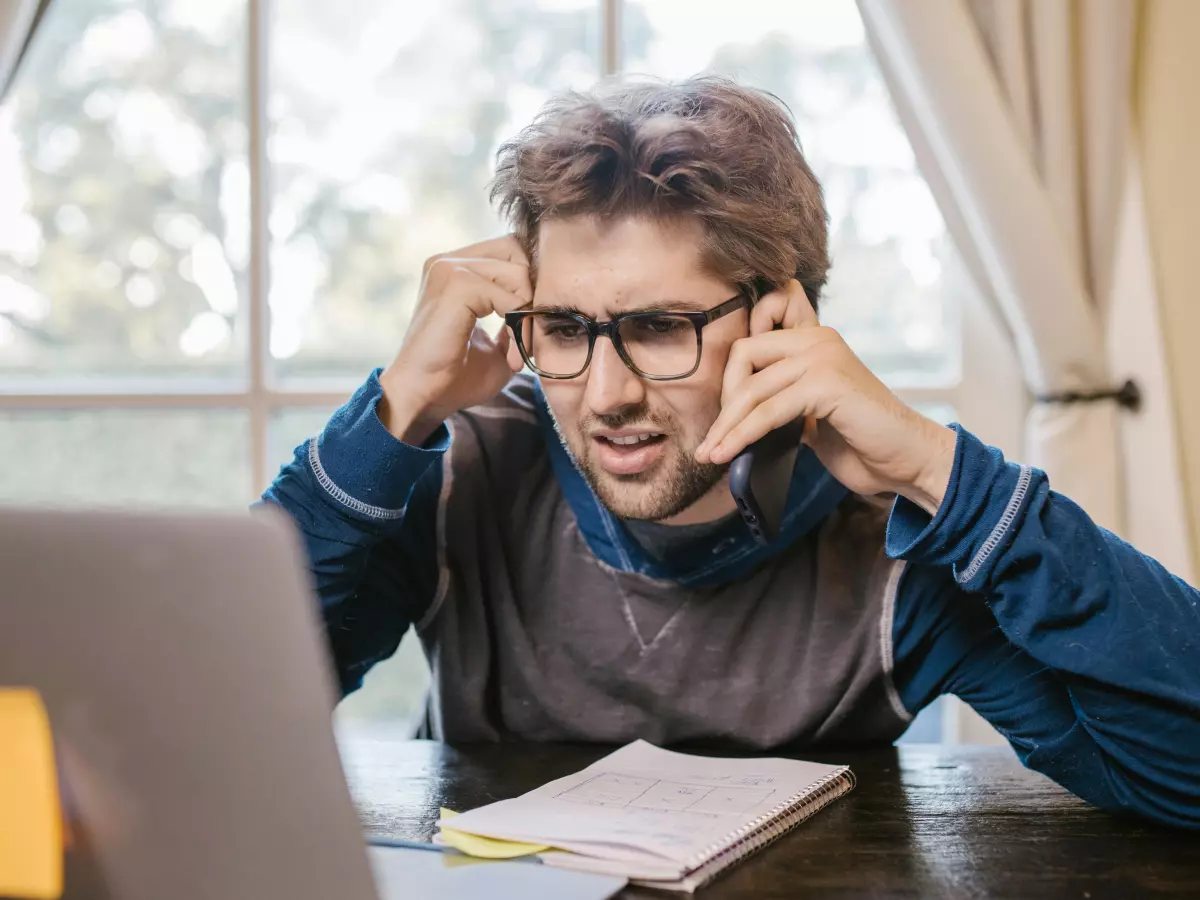 A man wearing glasses sits at a desk, looking at a laptop with a frustrated expression.