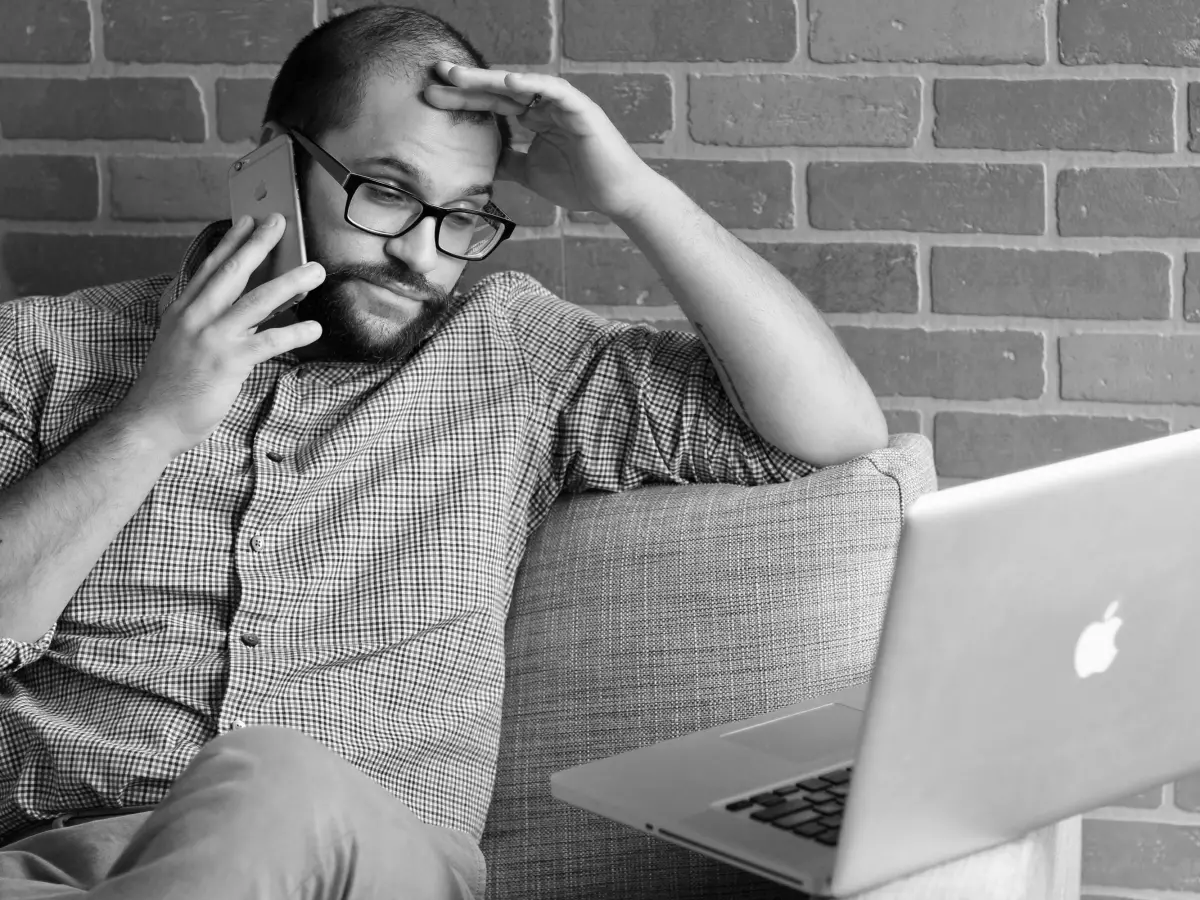 A man is sitting on a couch, looking frustrated while holding a phone to his ear. He's leaning back on the couch, with one hand on his forehead and the other holding the phone. He's wearing glasses and a checked shirt. A laptop is on the table next to him. The background is a brick wall.