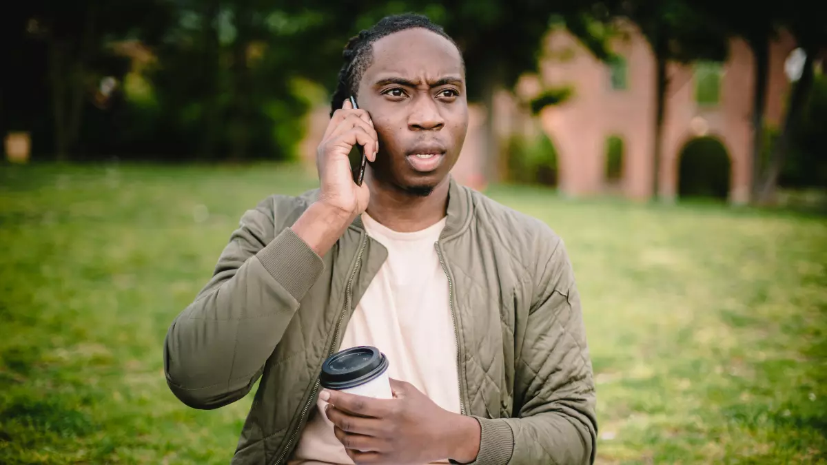 A man in a green jacket sits on a grassy lawn, talking on his phone and holding a coffee cup. He is looking to the side, seemingly engaged in conversation.