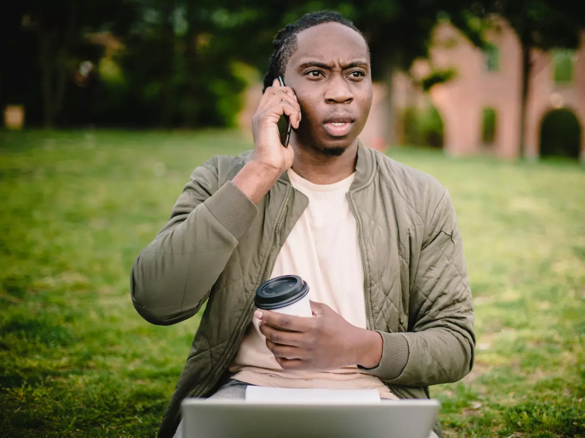 A man in a green jacket sits on a grassy lawn, talking on his phone and holding a coffee cup. He is looking to the side, seemingly engaged in conversation.