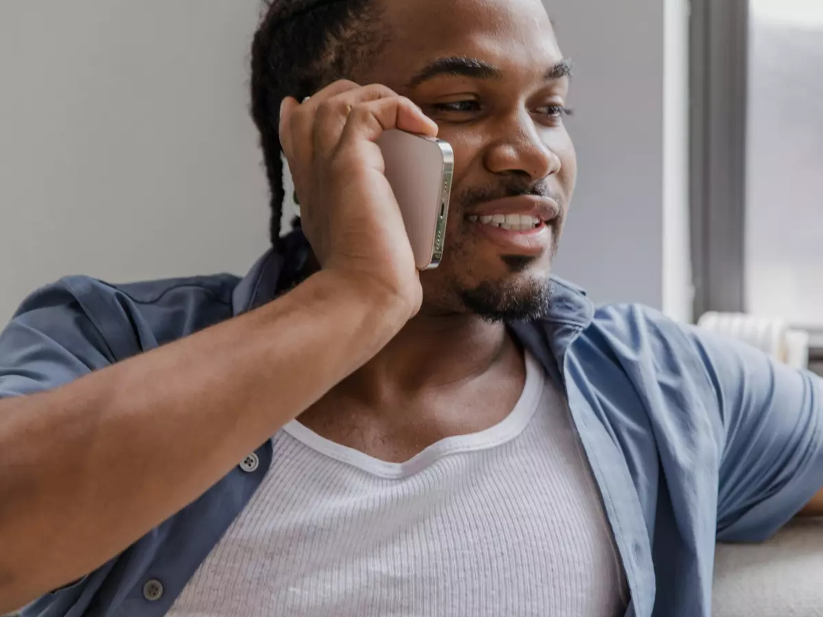 A young man with a beard is talking on his phone in front of a window.