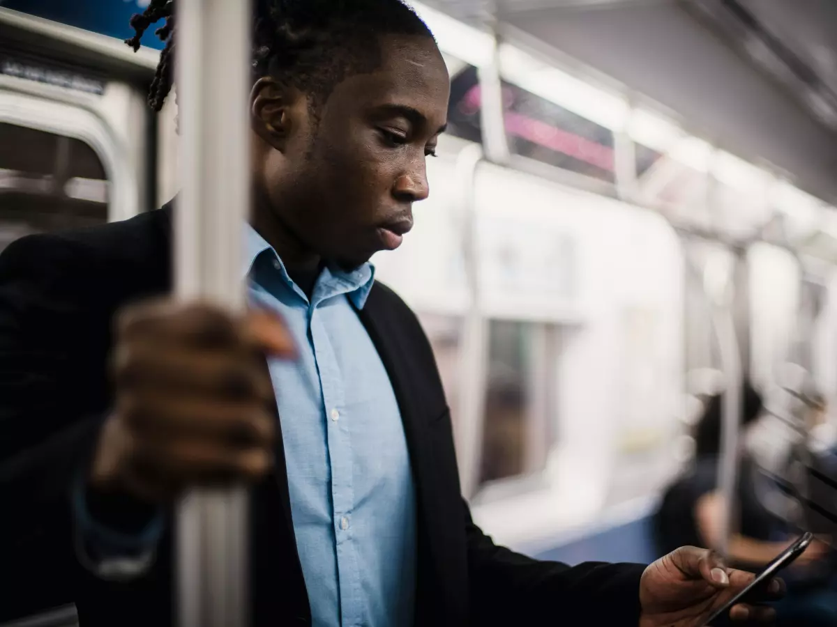 A man in a suit and tie is on a subway train, holding a smartphone in his hand and looking down at it.