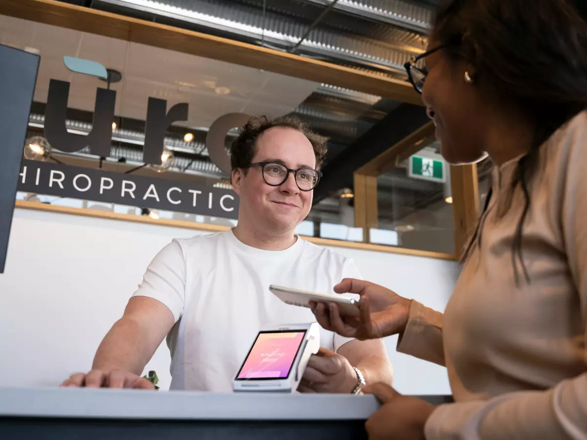 A man in a white shirt and glasses is standing at a counter in a chiropractic office, looking at a small device in his hand and smiling at a woman who is standing in front of him, also smiling.