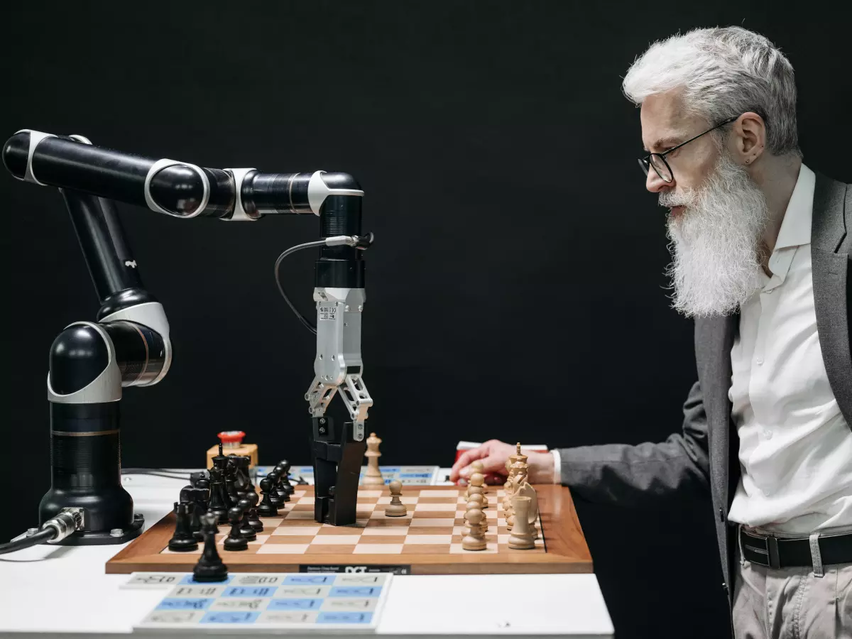 An elderly man plays chess against a robot arm. He is looking at the board with a serious expression, while the robot arm is reaching out to make a move.