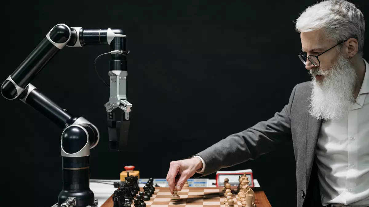 A robotic arm playing chess against an older man with a long white beard.