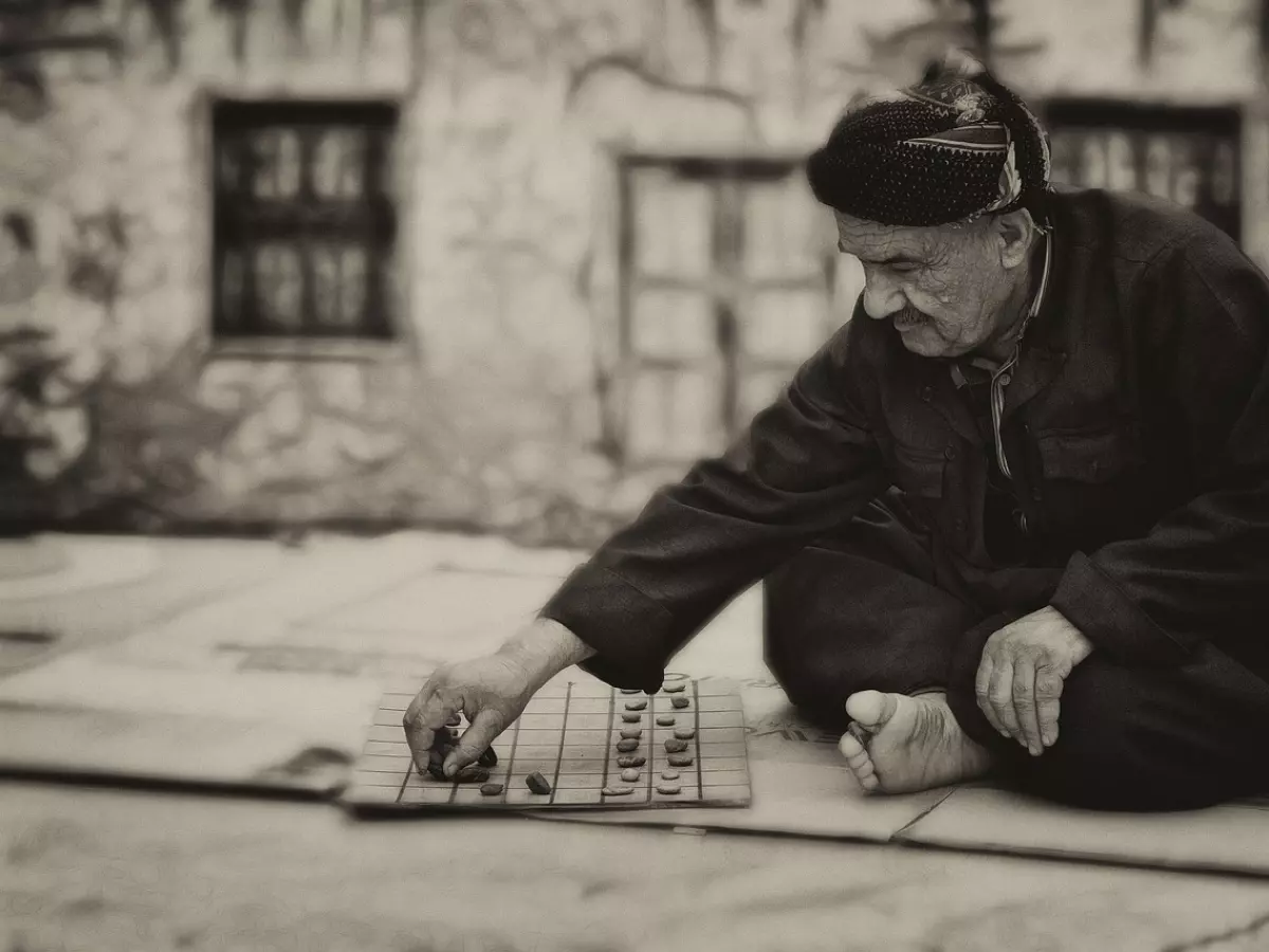 An elderly man sits on the ground, playing checkers with a focused expression.