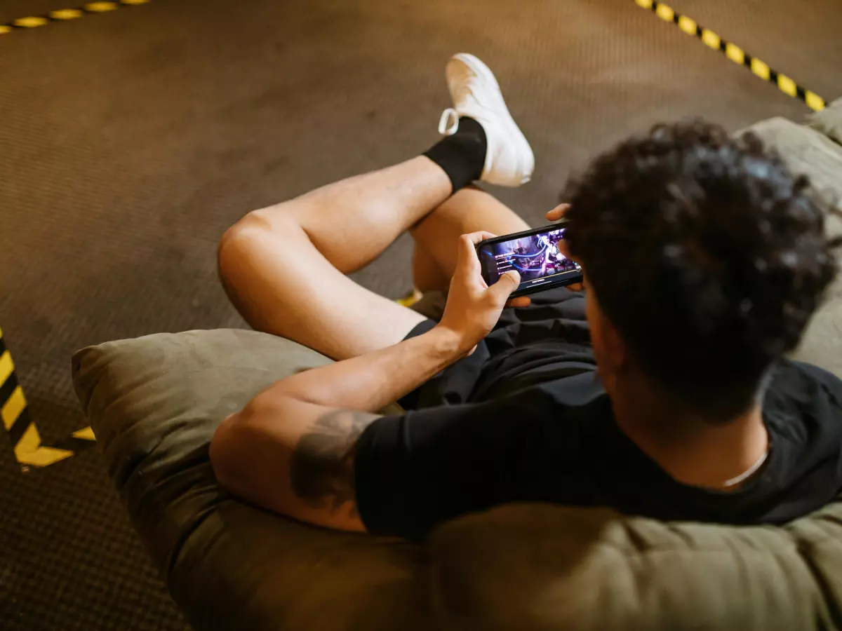 A young man sits on a couch and plays a video game on his smartphone.