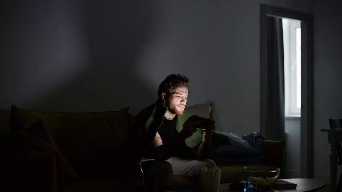 A person sitting on a couch in a dimly lit room, holding a smartphone in their hands. The phone is illuminated, highlighting its screen. The person