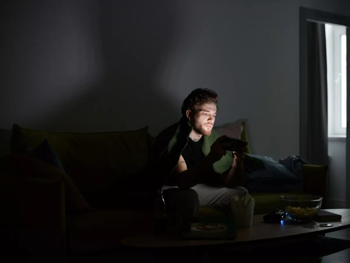 A person sitting on a couch in a dimly lit room, holding a smartphone in their hands. The phone is illuminated, highlighting its screen. The person's face is partially obscured by the phone.