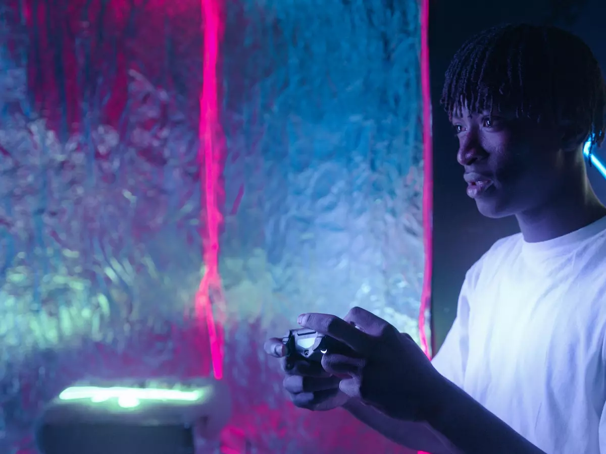 A man holds a gaming controller in a dark room with neon lights in the background.