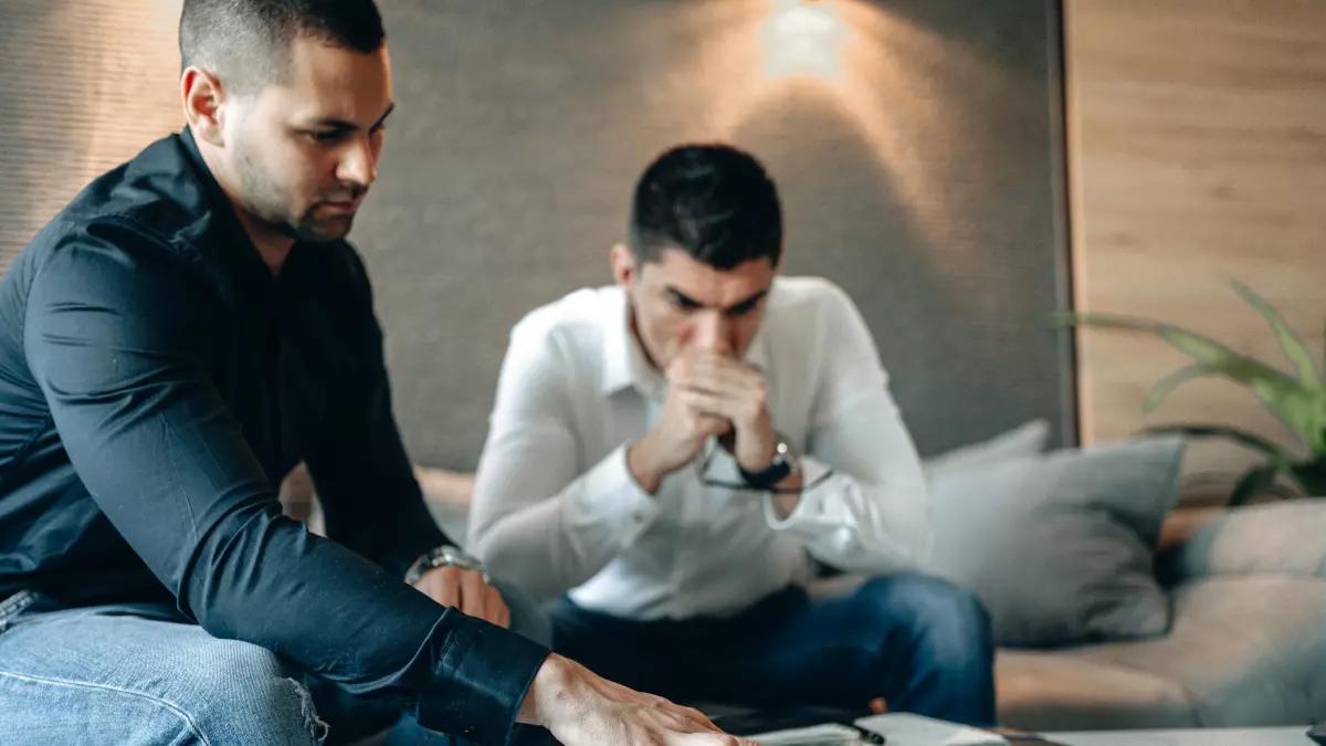 Two men sit on a couch in a living room, looking at a tablet, likely analyzing financial data. 