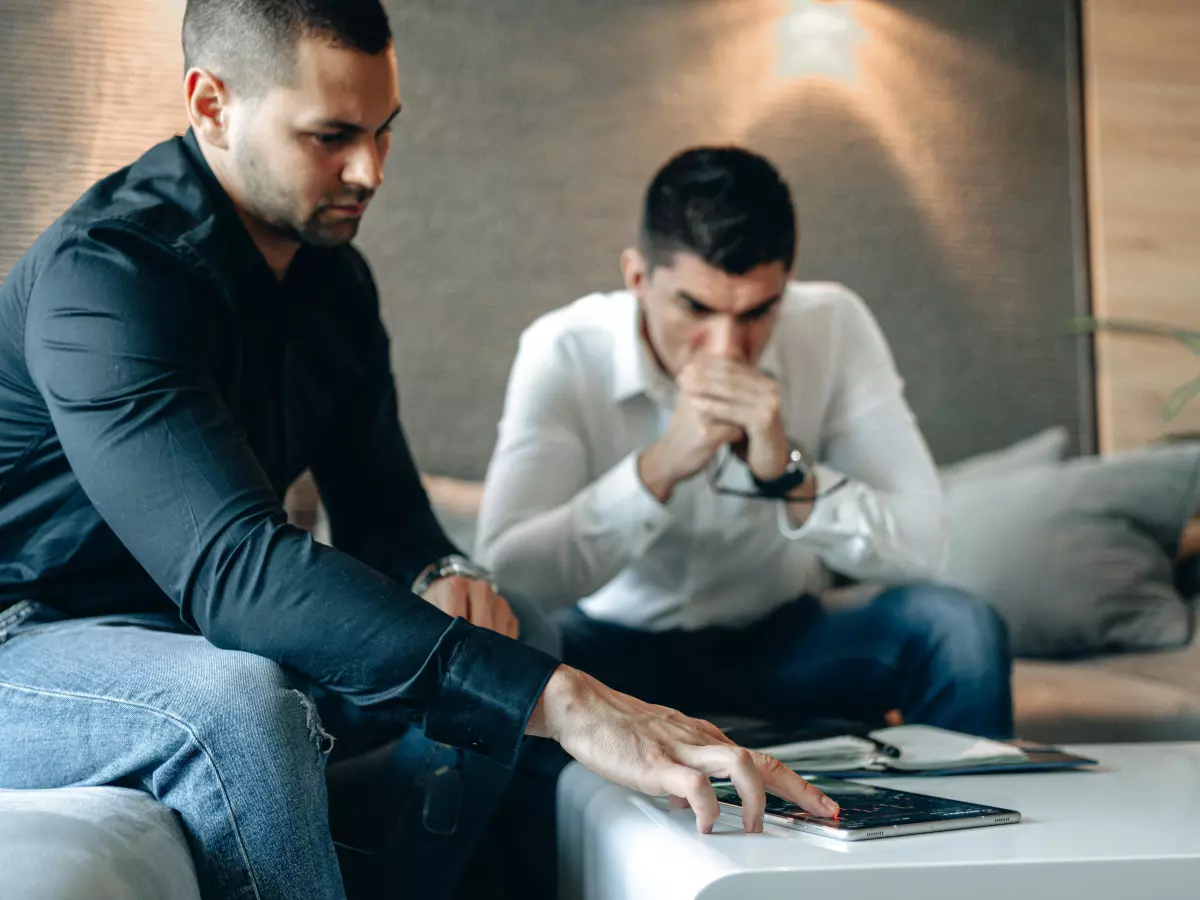 Two men sit on a couch in a living room, looking at a tablet, likely analyzing financial data. 