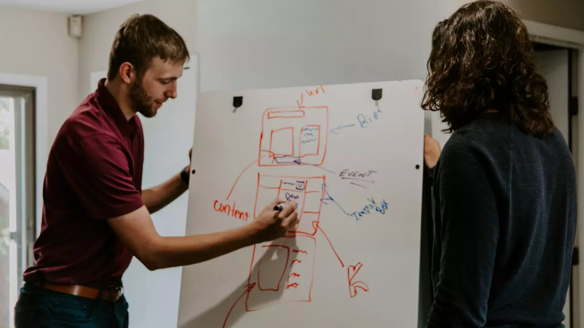 Two people working together on a whiteboard, one drawing a website layout and the other pointing to the content