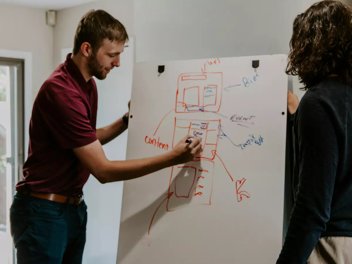 Two people working together on a whiteboard, one drawing a website layout and the other pointing to the content