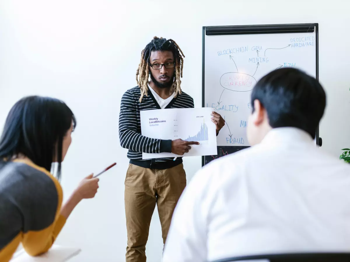 A young man, possibly an educator, stands in front of a whiteboard explaining a concept to a group of students.