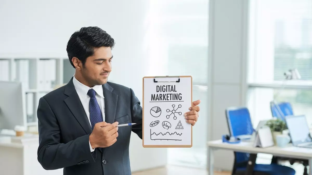 A businessman is standing in an office environment and holding a clipboard with a drawing of a digital marketing plan. He is using a pen to point at a specific element of the plan and looking directly at the camera. The background is blurred and depicts an office setting with computers and a desk. 