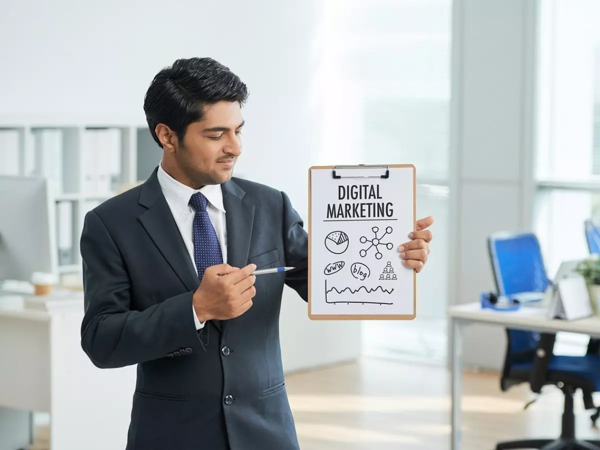 A businessman is standing in an office environment and holding a clipboard with a drawing of a digital marketing plan. He is using a pen to point at a specific element of the plan and looking directly at the camera. The background is blurred and depicts an office setting with computers and a desk. 