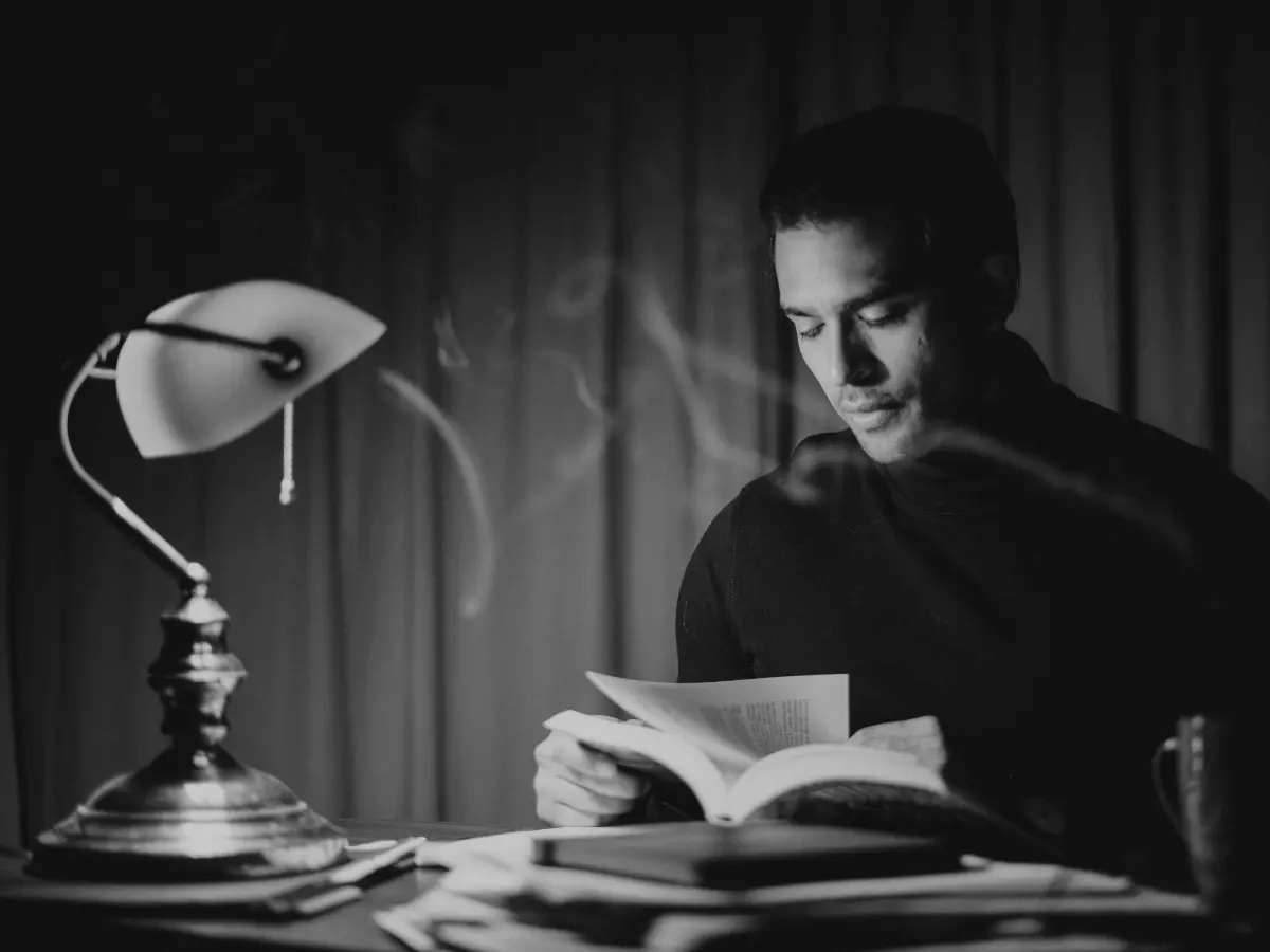 A man sitting at a desk in a dimly lit room, reading a book under a lamp. He is wearing a dark sweater and has a serious expression on his face.