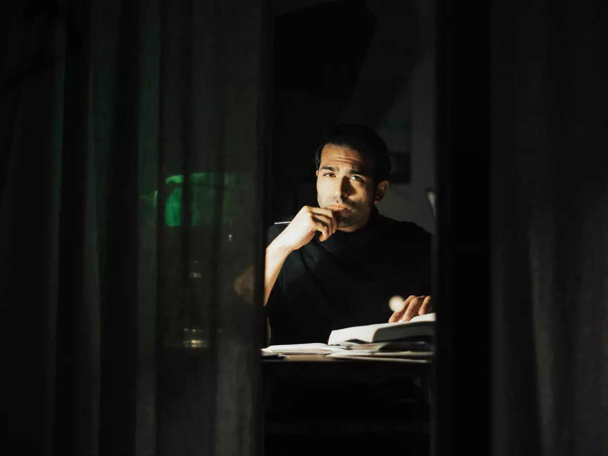 A man sits at a desk in a dimly lit room, reading a book. He is wearing a black shirt and has his chin resting on his hand. The image is taken from behind a curtain, creating a sense of mystery.