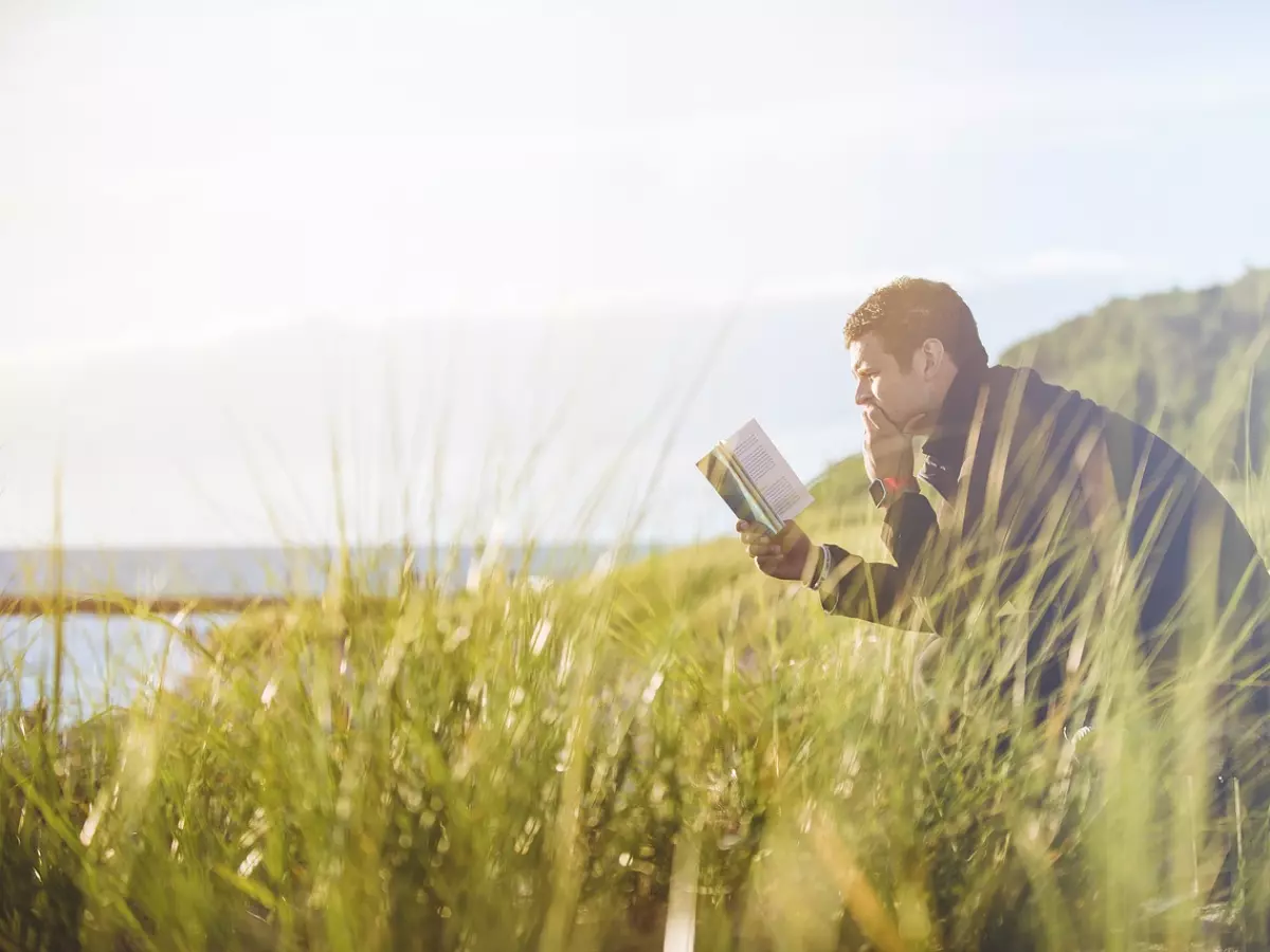 A man is sitting on a bench by the ocean and reading a book. The sun is shining brightly and the grass is green.