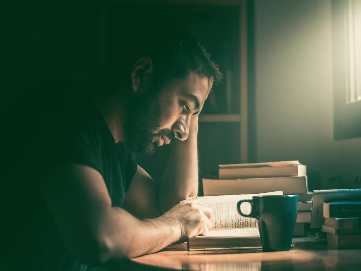 A man is sitting at a table, reading a book with a pen in hand. He has a serious expression on his face and is leaning forward, focused on the text.