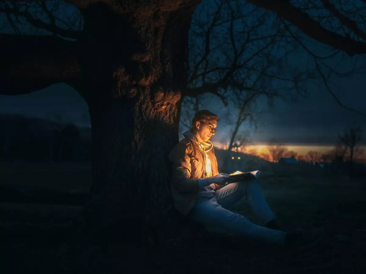 A person reading a book in the woods at night. There is a tree and a small house in the background. The image is lit by a soft light coming from the book.