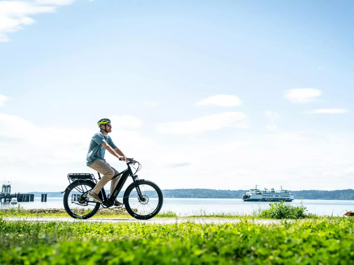 A man in a white shirt and blue jeans rides an electric bike on a path near a body of water.