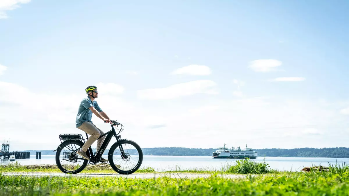 A man in a white shirt and blue jeans rides an electric bike on a path near a body of water.