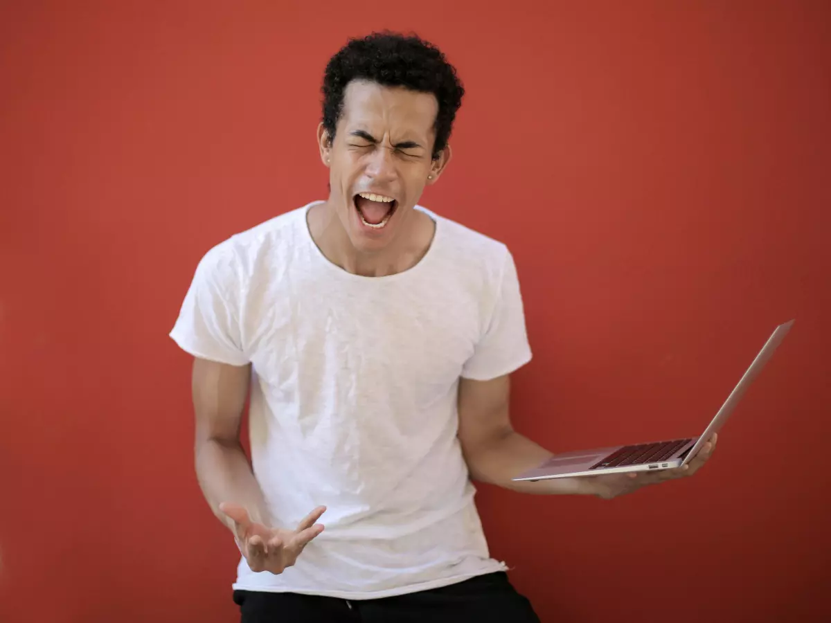 A man in a white T-shirt, standing in front of a red wall, screaming in frustration. He is holding a laptop in his hands.