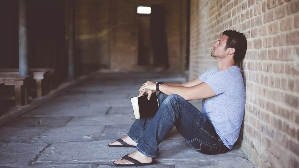 A man sitting on the floor with his back against a brick wall, holding a book in his lap. He is wearing a gray shirt and blue jeans. The photo is taken from a low angle. 